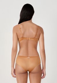 Donna vista di spalle che indossa un reggiseno beige abbinato con spalline regolabili e perizoma a vita alta, su uno sfondo chiaro e semplice.