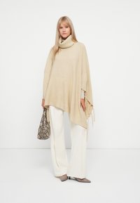 Poncho bege com riscas e um colarinho alto, apresentando bordas com franjas. Combinado com calças brancas de perna larga e uma mala texturizada bege.
