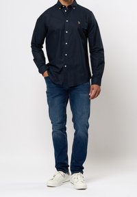 Granatowa koszula na długi rękaw z guzikami w kolorze białym, z haftowanym logo na piersi. Połączona z niebieskimi dżinsami i białymi trampkami.