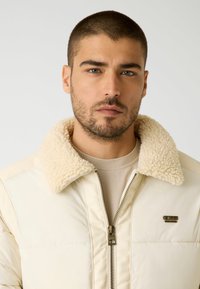 Giacca puffa color crema con un morbido colletto in shearling beige, chiusura con zip e dettaglio in metallo con logo sul petto sinistro. Texture liscia.