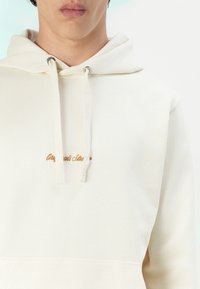 Crème kleurige hoodie met trekkoorden en zilveren oogjes. Beschikt over geborduurde tekst in bruin op de borst. Zachte stof en een voorzak.