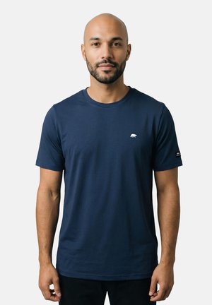 Mann mit Bart, der ein marineblaues kurzärmeliges T-Shirt mit kleinem weißem Bärenlogo auf Brust und Ärmel trägt, steht vor einfachem Hintergrund.