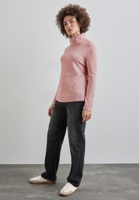Suéter de cuello alto rosa claro con un dobladillo grueso acanalado y dos botones decorativos, combinado con jeans oscuros y zapatillas blancas sobre un fondo gris.