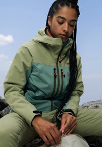 Giacca leggera e impermeabile con un design verde e teal, tasche con zip e accessori arancioni a contrasto. Texture liscia e vestibilità ergonomica.