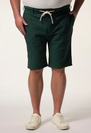 Mand iført mørkegrønne snøreshorts med lommer, hvide sneakers og en hvid skjorte med hænderne i lommerne.