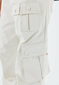 Pantalones cargo color crema hechos de un tejido ligero, con grandes bolsillos con tapa y cierres de botón, y una textura suave.