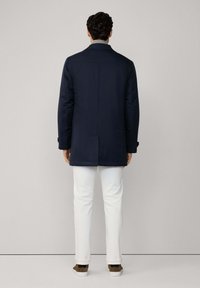 Marineblauwe middelgrote jas met een gladde textuur, enkele split en een puntkraag. Gecombineerd met witte broek en bruine lage schoenen.