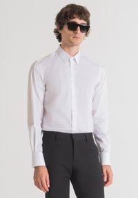 Camicia bianca a maniche lunghe con colletto a punta e chiusura con bottoni. Indossata con pantaloni neri su misura e occhiali da sole scuri. Tessuto liscio, senza motivi.