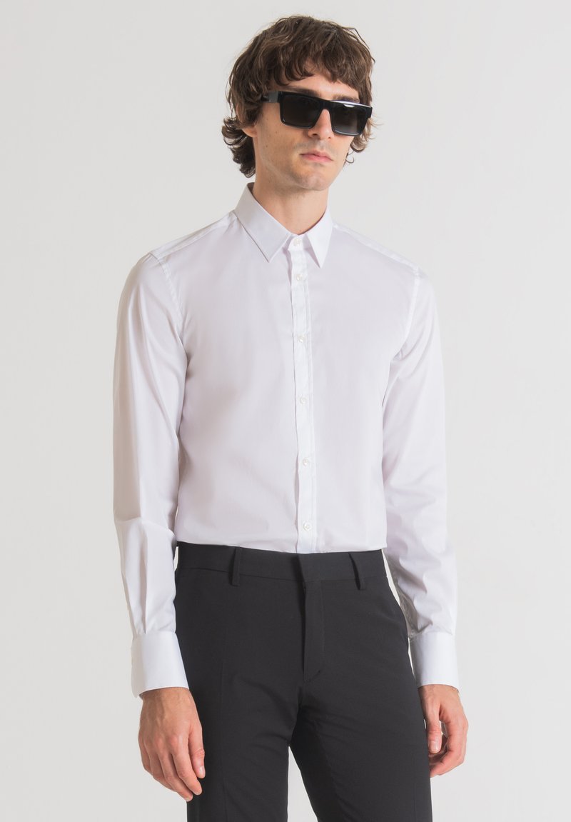 Camicia bianca a maniche lunghe con colletto a punta e chiusura con bottoni. Indossata con pantaloni neri su misura e occhiali da sole scuri. Tessuto liscio, senza motivi.