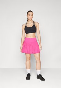 Sujetador deportivo negro con tirantes ajustables, combinado con una falda plisada rosa brillante. Usado con zapatos negros y calcetines tobilleros blancos.