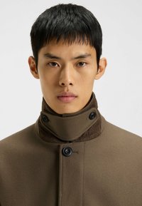 Mand med kort, sort hår iført en højhalset, brun frakke, der er knappet ved halsen og brystet, som står fremadvendt mod en ensfarvet baggrund.