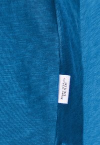 Modrá džínová látka s žebrovanou strukturou, s viditelnou bílou etiketou s nápisem "Marc O'Polo DENIM." Hladké okraje a strukturovaný tvar.