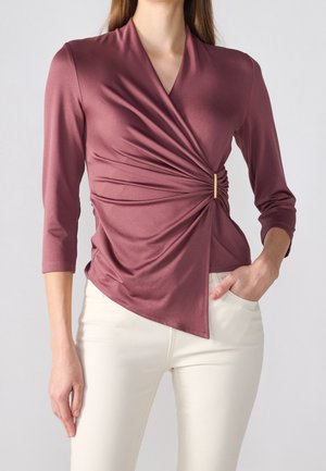 Femme portant un haut cache-cœur mauve avec des manches trois-quarts et un ourlet asymétrique, associé à un pantalon ajusté écru.