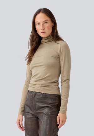 Beige langärmliges Rollkragenoberteil aus glattem, dehnbarem Stoff. Kombiniert mit enganliegenden, dunkelbraunen Lederhosen.