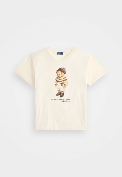 Kremkrāsas īsās piedurknes T-krekls ar grafiku, uz kuras ir lācis ziemas apģērba, brūnu akcentu un uzraksts "POLO BEAR by RALPH LAUREN."