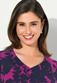 Kobieta z włosami do ramion, w brązowym kolorze, uśmiecha się, nosząc fioletową bluzkę w kwiatowy wzór z dekoltem w serek oraz srebrne kolczyki w kształcie liści.