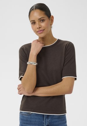 Kobieta o ciemnych włosach związanych do tyłu, ubrana w brązową bluzkę z krótkim rękawem i białą lamówką oraz niebieskie dżinsy, opierająca podbródek na dłoni z srebrną bransoletką.