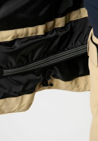 Schwarze und beige Jacke mit Innenfutter, das einen Reißverschluss, eine verstellbare Schnur und eine glatte Textur mit kontrastierenden horizontalen Streifen aufweist.