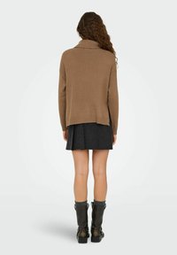 Un pull oversized marron avec un col haut, associé à une jupe plissée noire et des bottes noires montantes avec des accents en tricot gris.