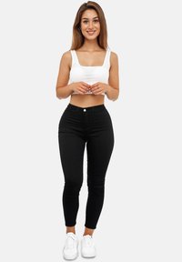 Mujer de pie sonriendo, vestida con un top corto blanco, jeans ajustados negros de cintura alta y zapatillas blancas, frente a un fondo sencillo.