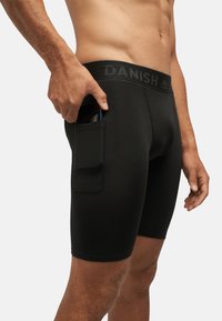 Boxer shorts neri con un design aderente, vita elasticizzata etichettata "DANISH" e una tasca laterale che contiene un dispositivo mobile.