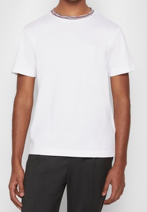Homme portant un T-shirt blanc uni à manches courtes avec un logo de marque discret sur la poitrine et un pantalon gris foncé.