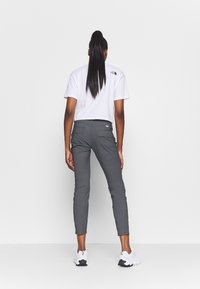 Kobieta z długim kucykiem, ubrana w białą krótką koszulkę, szare dopasowane spodnie i białe sneakersy, stojąca na jednolitym białym tle.