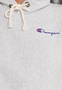 Light grey hoodie with a drawstring closure and metal eyelets. Features a blue embroidered "Champion" logo on the front. Soft texture.