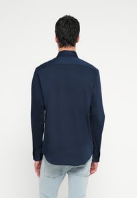 Tumesinise värvusega pika varrukaga särk, millel on struktureeritud krae, sile tekstuur ja nööpidega mansetid. Tagantvaates on näha kaardus ääred ja minimaalne detail.