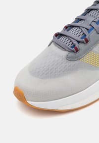 Pilkas sportinis batelis su tinklo viršutine dalimi, tekstūruota apdaila, geltonais akcentais ir oranžiniu padu. Įrengtas tradicinis raištelių užsegimas.