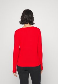 Pull rouge à manches longues avec encolure ronde et poignets côtelés, en matière tricotée douce, cintré à la taille, vu de dos.