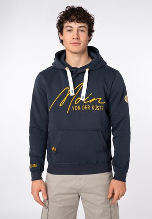 Jonge man met krullend haar die een donkerblauwe hoodie met gele tekst "Moin von der Küste" en beige broek draagt, staand tegen een effen achtergrond.