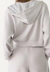 Gekürzter Kapuzenpullover aus hellgrauem Stoff mit Kapuze, weicher Textur, geripptem Saum und langen Ärmeln, kombiniert mit passenden Hosen.