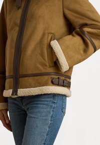 Giacca in shearling marrone con un esterno liscio, rivestimento crema morbido, chiusura a zip, dettagli in pelle e cinghie regolabili in vita.