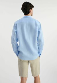 Jasnoniebieska koszula z długim rękawem o luźnym kroju, z karczkiem z tyłu i okrągłym dołem, zestawiona z beżowymi szortami. Gładki, lekki materiał.