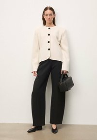 Veste blanche sur mesure avec boutons noirs, associée à un pantalon noir à jambes larges et un sac à main noir. Texture lisse, design structuré.