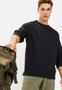 Donkerblauwe sweatshirt met raglanmouwen, geribbelde manchetten en een ronde halslijn. Gecombineerd met olijfgroene broek en een kaki rugzak.