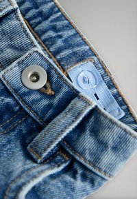 Jeansabteilung mit hellblauem Stoff, silbernem Metallknopf, kontrastierender Naht und einem angenähten Etikett mit Knopfloch.