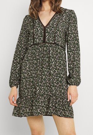Kvinde iført en sort blomsterkjole med lange puffede ærmer, V-halsudskæring og blondekantdetaljer, stående mod en ensfarvet lys baggrund.