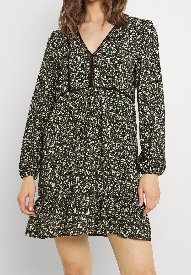 Femme portant une robe noire à fleurs avec de longues manches bouffantes, un décolleté en V et des détails en dentelle, debout devant un fond clair uni.