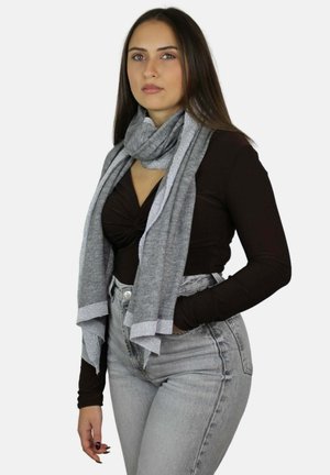 Giovane donna con capelli castani lunghi che indossa una maglietta nera, jeans grigio chiaro e una sciarpa grigia su uno sfondo semplice.