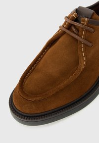 Zapato de ante marrón con cordones, con costuras en contraste y suela negra, mostrando un primer plano detallado de la puntera y los cordones.