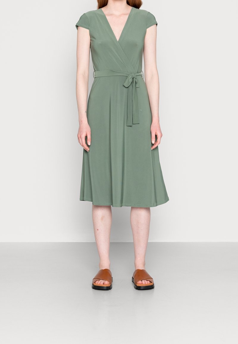 Femme portant une robe portefeuille olive longueur genou avec manches courtes et ceinture à la taille, associée à des sandales ouvertes marron.