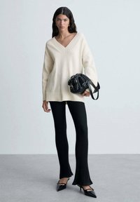 Pulover crem cu decolteu în V, pantaloni negri cu model în relief, geantă neagră cu accente aurii și pantofi negri cu vârf ascuțit. Textură netedă, croială relaxată, siluetă stratificată.