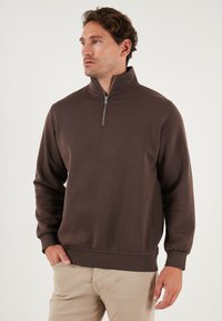 Bruine rits sweatshirt met lange mouwen, hoge kraag, geribde boorden en een zachte textuur, gecombineerd met lichtgekleurde broek.