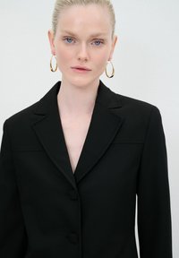 Blazer nero con colletto a revers, due bottoni frontali e dettagli di cucitura sottili. La modella indossa orecchini a cerchio dorati. Tessuto dalla superficie liscia.