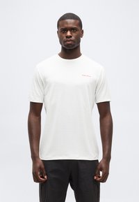 Άνδρας που φοράει απλό λευκό κοντομάνικο μπλουζάκι με μικρό κόκκινο λογότυπο στο στήθος, συνδυασμένο με μαύρο παντελόνι, στέκεται μπροστά σε απλό φωτεινό φόντο.