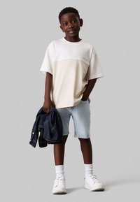 Camiseta color bloque en crema y beige con mangas cortas, combinada con pantalones cortos de mezclilla azul claro. Zapatillas blancas y una chaqueta de mezclilla oscura.