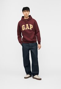Jeune homme portant un sweat à capuche GAP bordeaux, un jean bleu foncé et des baskets blanches, debout avec une main dans la poche devant un fond uni.