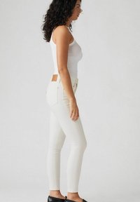 Jeans ajustados blancos con una textura suave, cintura alta y bolsillos traseros. Presenta un parche de cuero en la cintura. Estilizados con una camiseta de tirantes blanca.